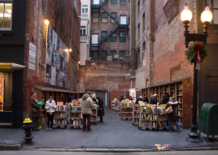 Librairie extérieure cosy dans une ruelle urbaine avec des gens feuilletant des livres entourés de murs de briques et de lampadaires vintage.
