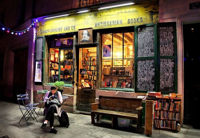 Librairie chaleureuse et grandiose la nuit avec un éclairage tamisé, remplie de livres et une personne lisant à l'extérieur.