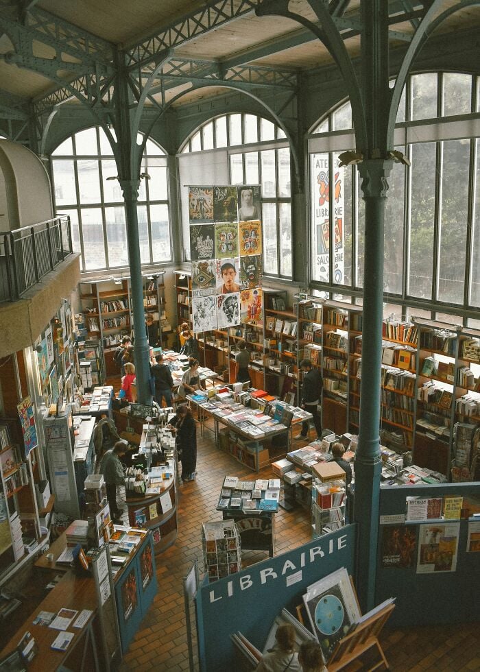Bibliothèque spacieuse et grandiose avec de hautes fenêtres cintrées, de hautes colonnes métalliques et des étagères remplies de livres et de visiteurs en train de les parcourir.