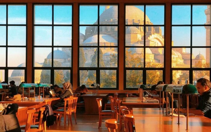 Acogedor interior de biblioteca con grandes ventanales que muestran una arquitectura grandiosa, y personas leyendo y estudiando en mesas de madera.