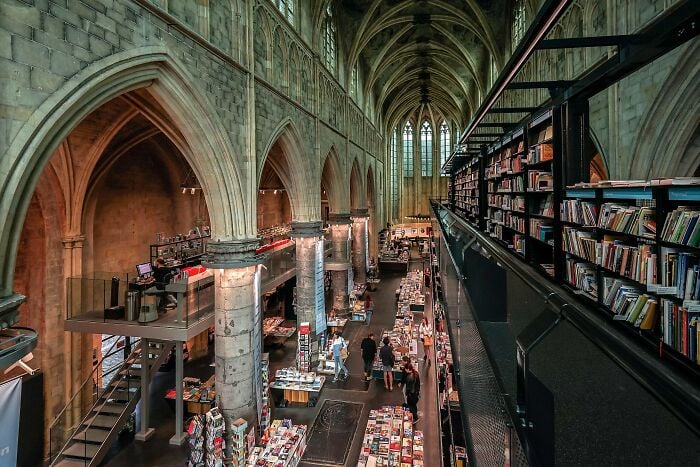 Grandioso interno di biblioteca con soffitti a volta, archi in pietra e file di scaffali pieni di libri e visitatori che curiosano.