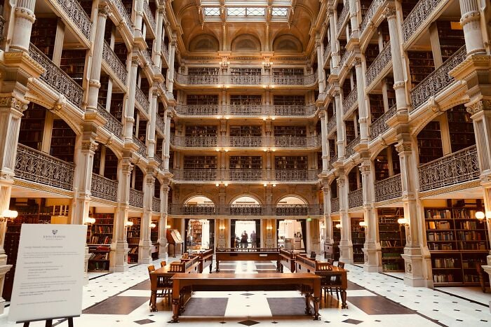 Interior de una grandiosa biblioteca con varios niveles de estanterías, columnas ornamentadas y una espaciosa zona de lectura con mesas y sillas de madera.
