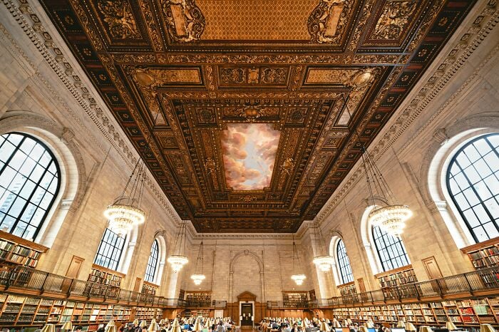 Interior de biblioteca grandioso con techo ornamentado, grandes ventanales arqueados, candelabros y filas de estanterías repletas de libros.