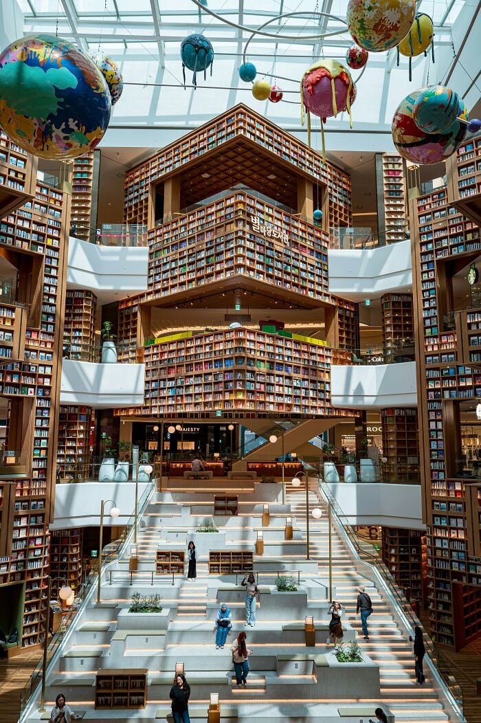 Interior de biblioteca grandioso con estanterías imponentes, gran claraboya y esculturas coloridas de planetas colgando del techo.
