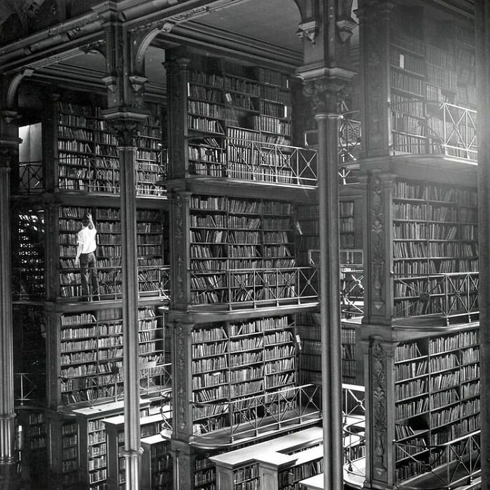 Grandiose library interior with towering bookshelves, multiple balconies, and a person reaching for a book in a magical setting.