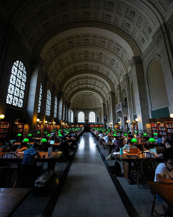 Grandiose library interior with high arched ceilings, large windows, and rows of study tables with green lamps and readers.