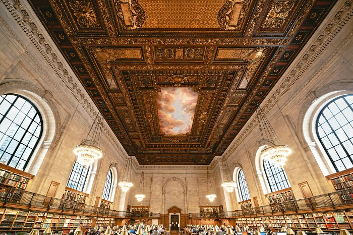 Grandiose library interior with ornate ceiling, large arched windows, chandeliers, and rows of bookshelves filled with books.