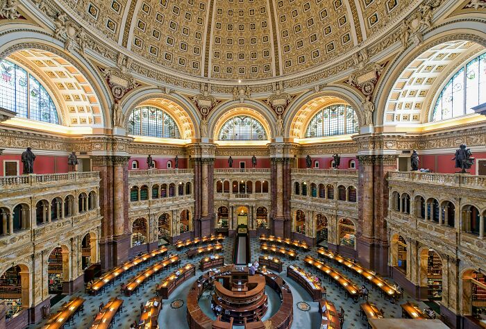 Grandiose and cosy library interior with ornate dome ceiling, arched windows, and rows of reading desks with warm lighting.